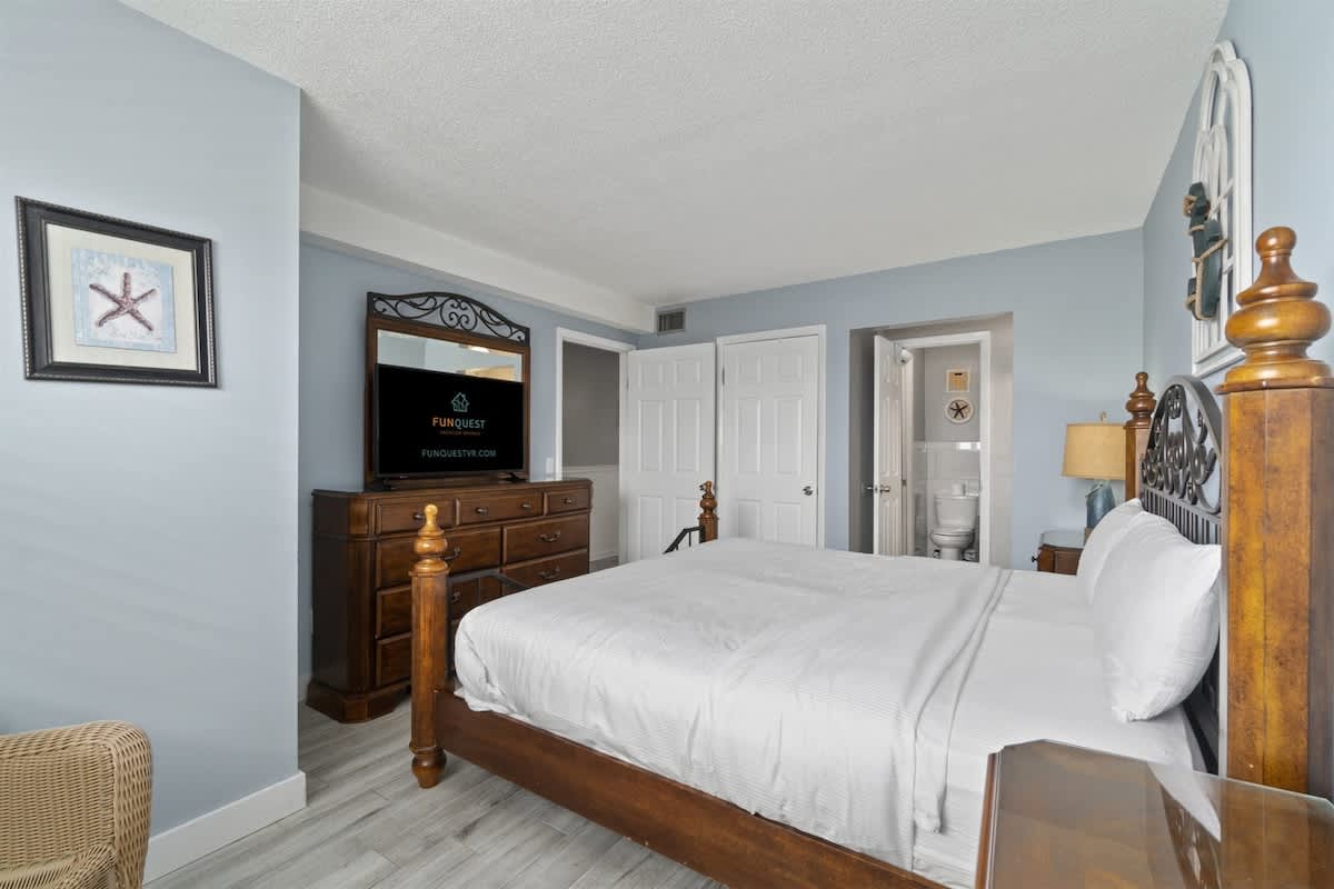 The Summit 728 – Bedroom with a wooden bed, dresser, TV, and bathroom door visible.