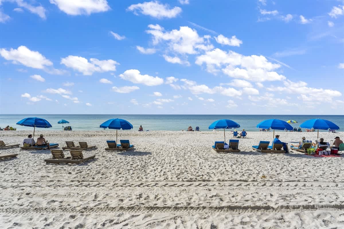 The Summit 728 – Beachfront view with blue umbrellas, lounge chairs, and people enjoying the sun on light-colored sand.