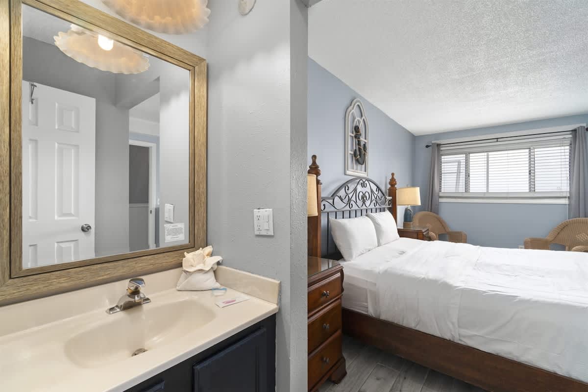 The Summit 728 – Bedroom with a bed, nightstand, two chairs, and bathroom mirror reflecting the sink area.