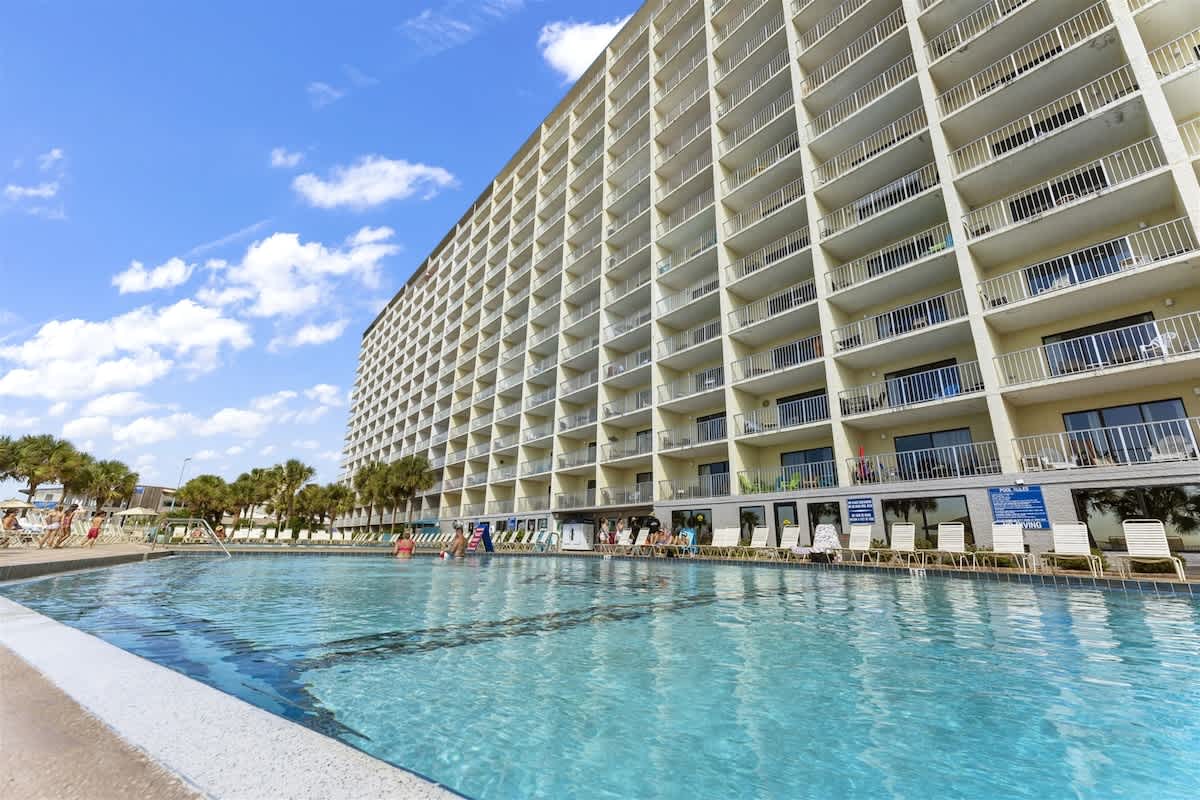 The Summit 728 – Poolside view of a tall building with balconies and palm trees under a blue sky.
