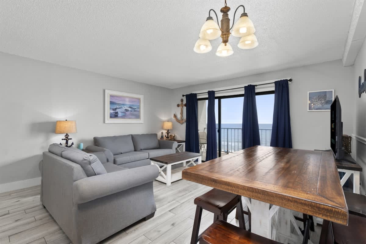 The Summit 728 – Living room with gray furniture, ocean view, and wooden dining table.