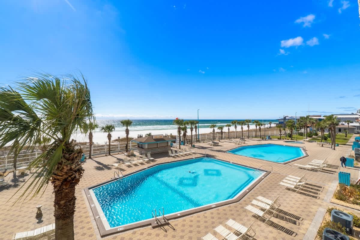 The Summit 1130 – View of two swimming pools, palm trees, and beach in the background under a clear blue sky.