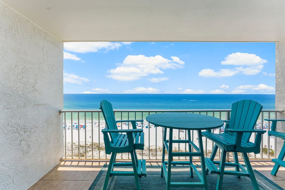 The Summit 1130 – Balcony with a round table and chairs, overlooking the beach and ocean under a blue sky.