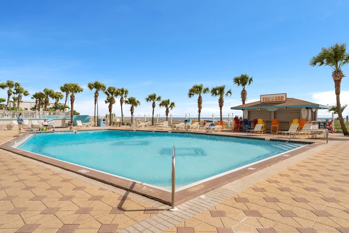 The Summit 1130 – Pool area with tiki bar, palm trees, and sun loungers against a clear blue sky.