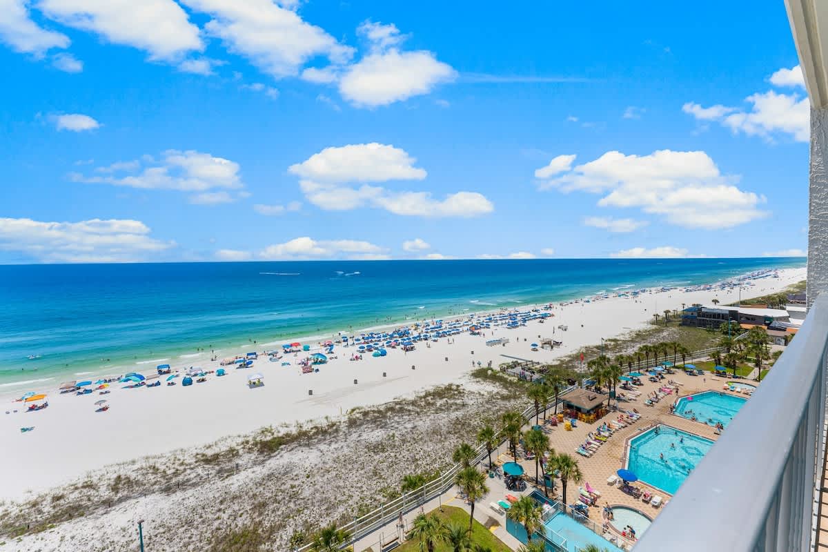 The Summit 1130 – Beachfront view from a balcony, showing sandy beach, colorful umbrellas, and swimming pools below.