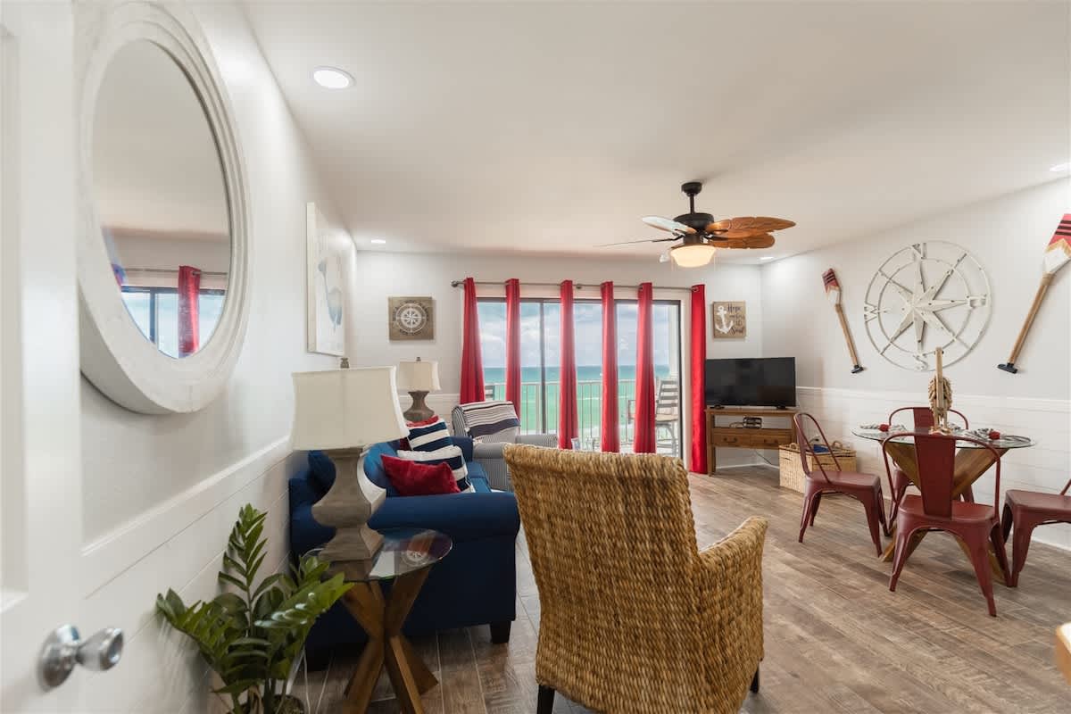 The Summit 504 – Living room with a sofa, chairs, dining table, TV, and ocean view through large windows with red curtains.