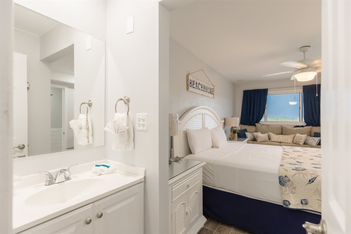 The Summit 504 – Bright bedroom with white furniture, large mirror, and cozy seating area by the window.