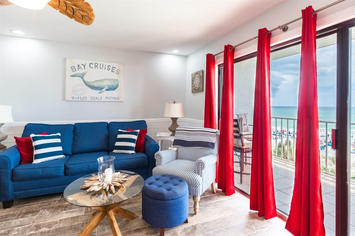 The Summit 504 – Living room with blue couch, striped chair, red curtains, and ocean view through glass doors.