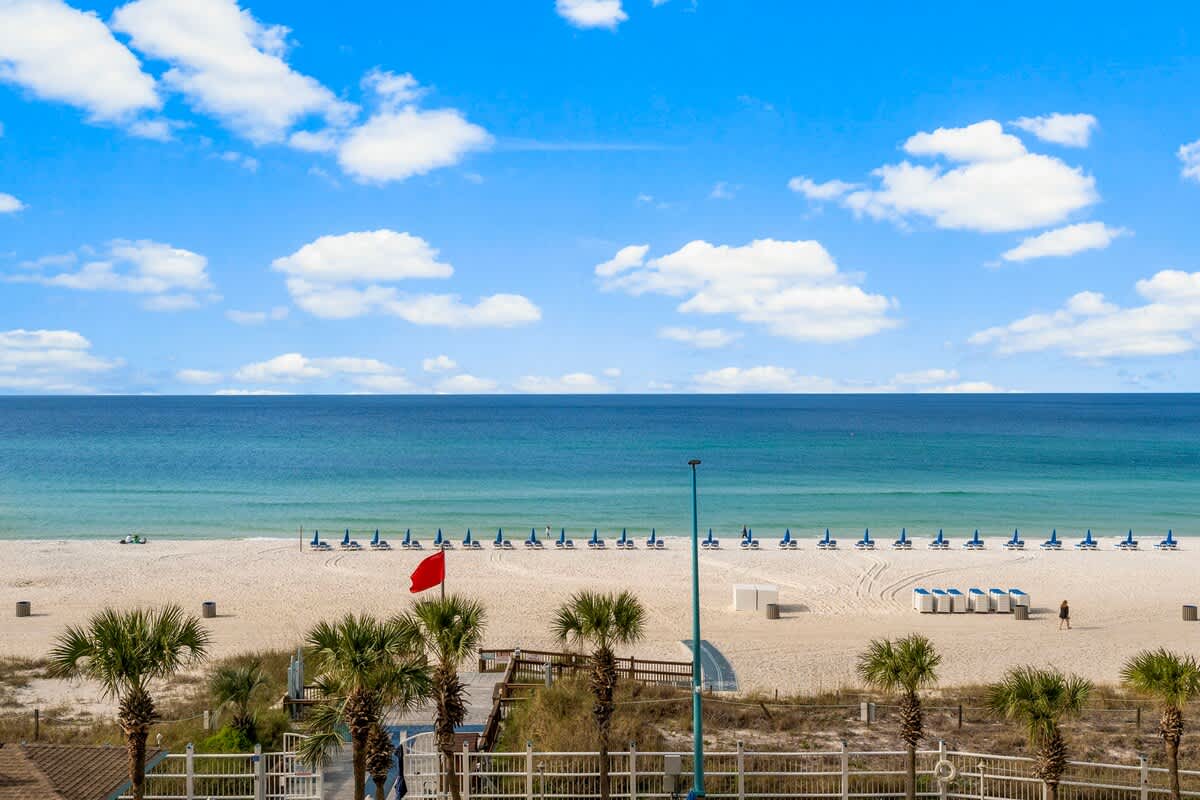The Summit 515 – Beach view with lounge chairs, palm trees, and ocean under a blue sky with clouds.