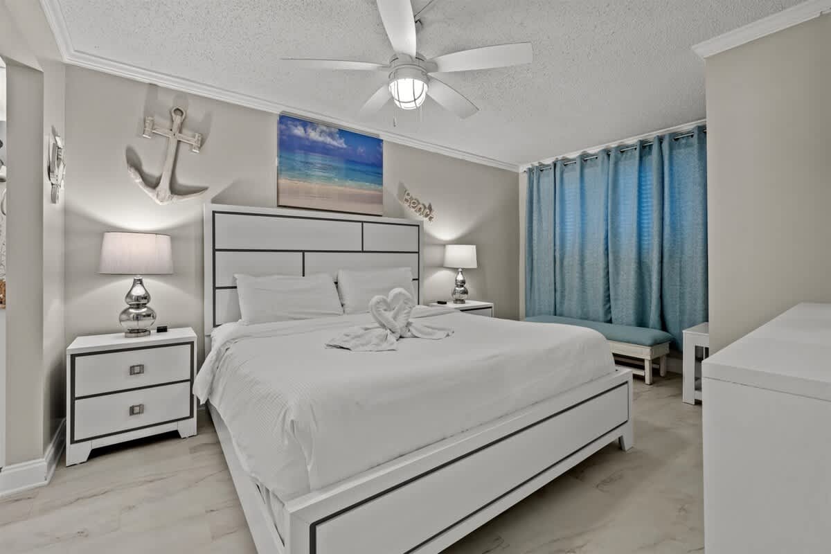 The Summit 816 – Modern bedroom with white furniture, a ceiling fan, and blue curtains. Features a decorative anchor and heart-shaped towels.