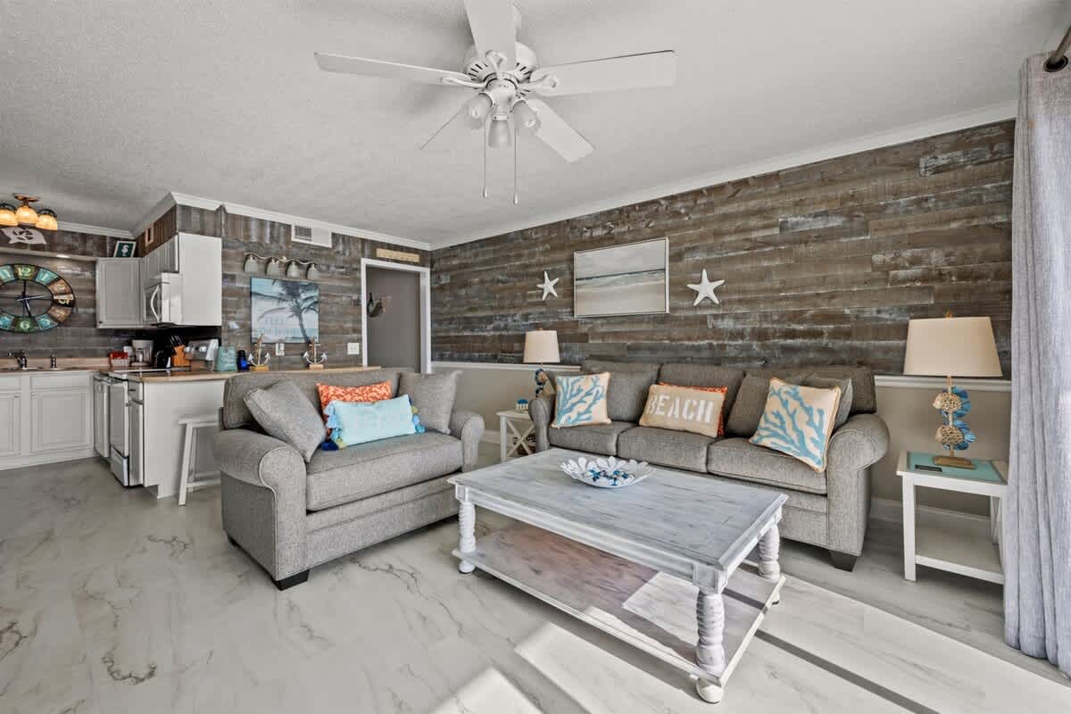 The Summit 816 – Living room with gray sofas, ocean-themed decor, wooden accent wall, and open kitchen concept.