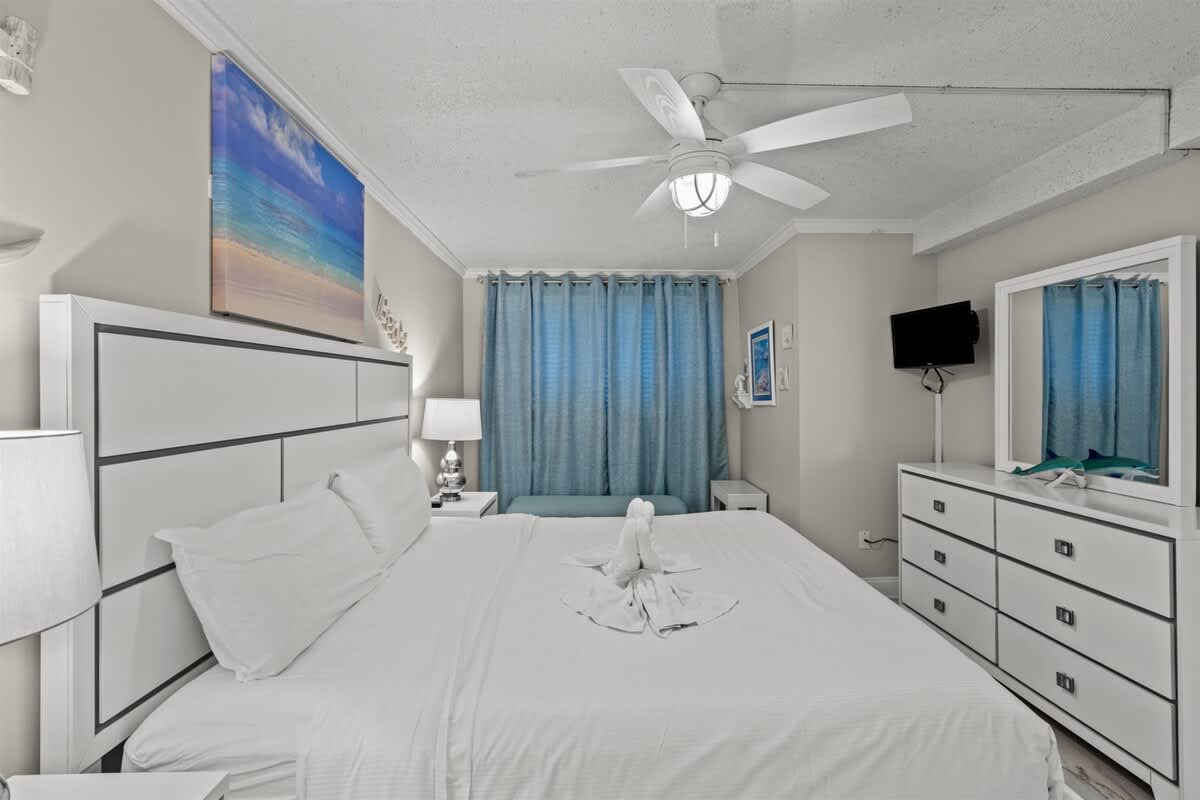 The Summit 816 – Bright bedroom with a large bed, blue curtains, dresser, TV, and decorative artwork.