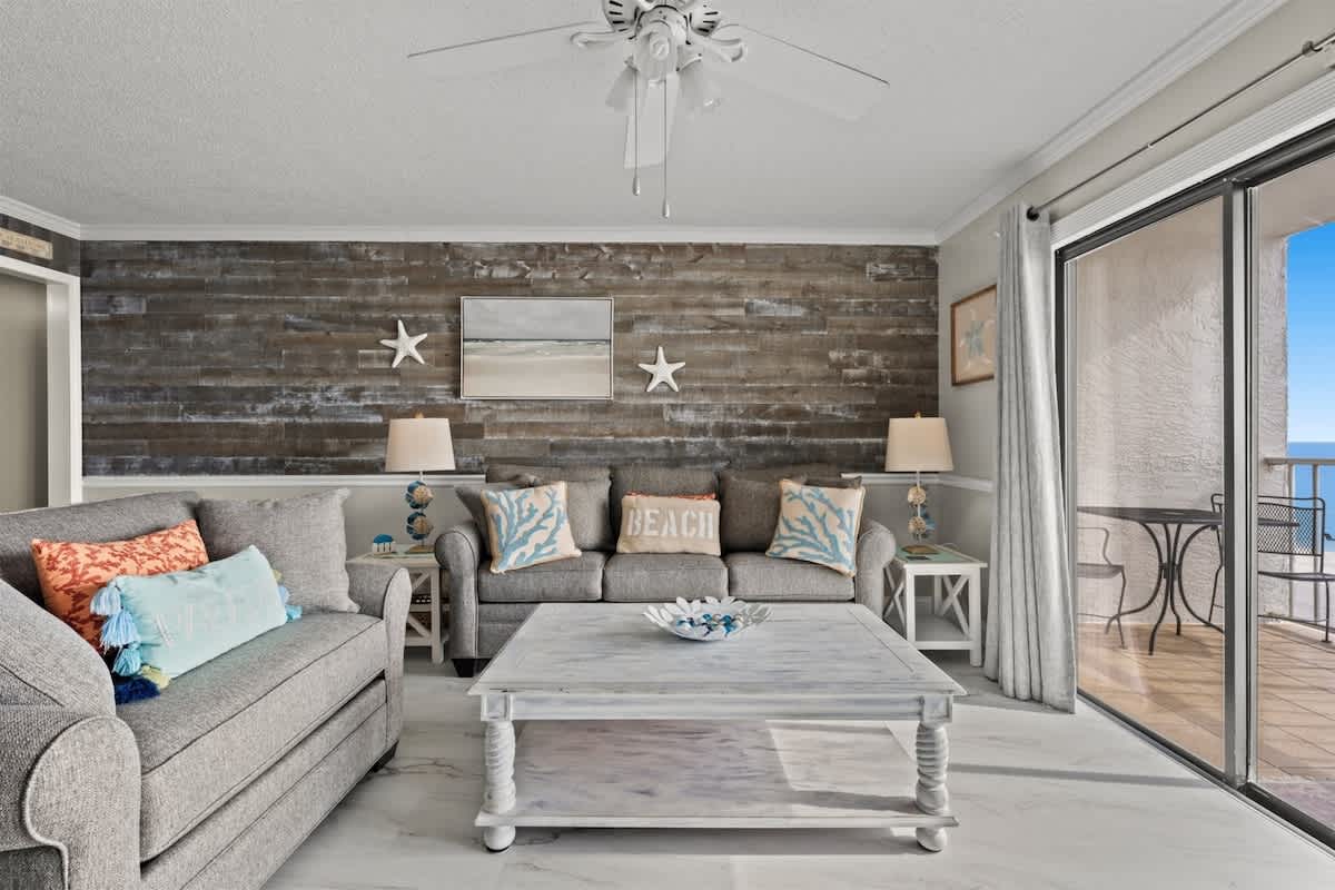 The Summit 816 – Living room with grey sofas, wooden accent wall, coffee table, and ocean view through sliding glass doors.