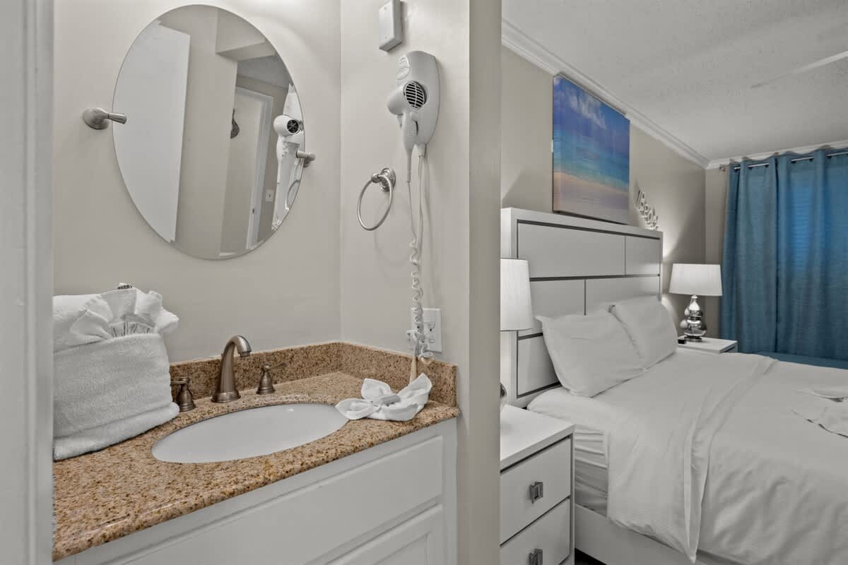 The Summit 816 – Bathroom sink area with mirror, hairdryer, and bedroom featuring a bed, bedside lamps, and blue curtains.