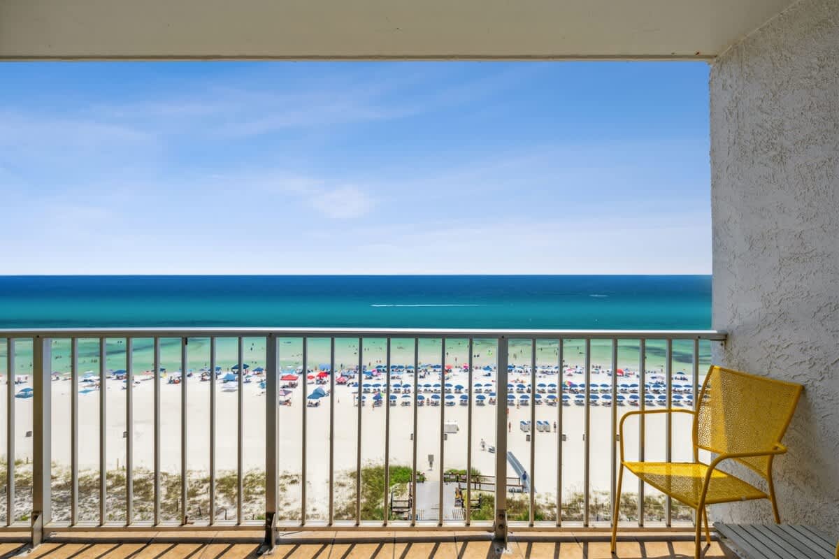 The Summit 1017 – Balcony view of beach, ocean, and yellow chair, with umbrellas on the sand.