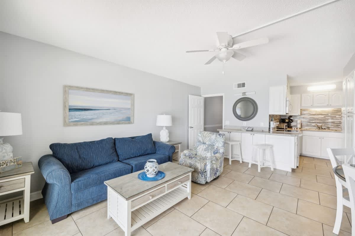 The Summit 1017 – Living room with blue couch, armchair, coffee table, and kitchenette featuring white cabinets and tile floor.