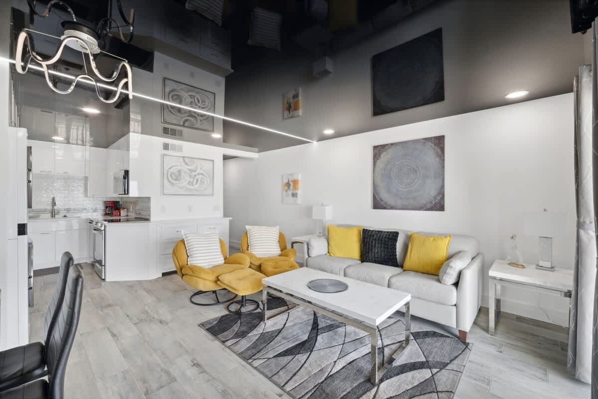 The Summit 702 – Modern living room featuring a white sofa, two yellow chairs, dining area, and kitchen with stainless appliances.
