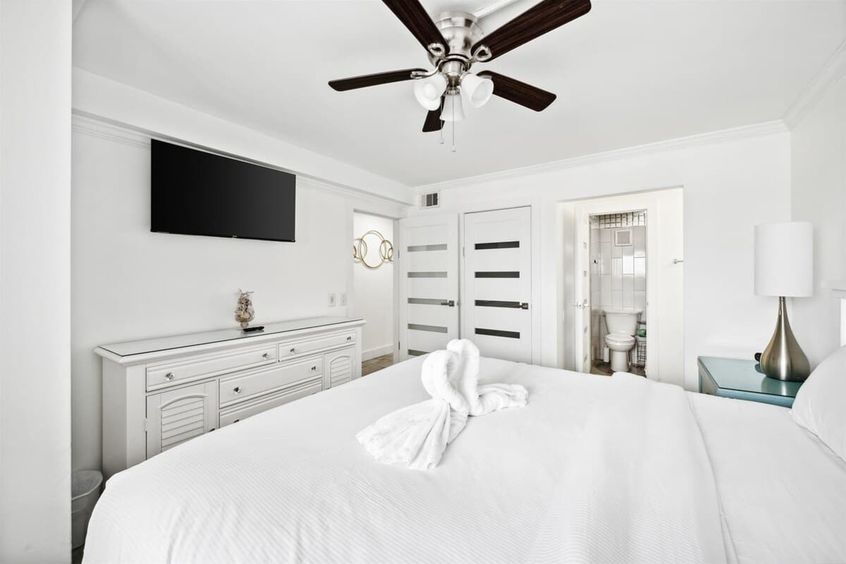 The Summit 702 – Bedroom with a white bed, ceiling fan, television, and bathroom door visible.