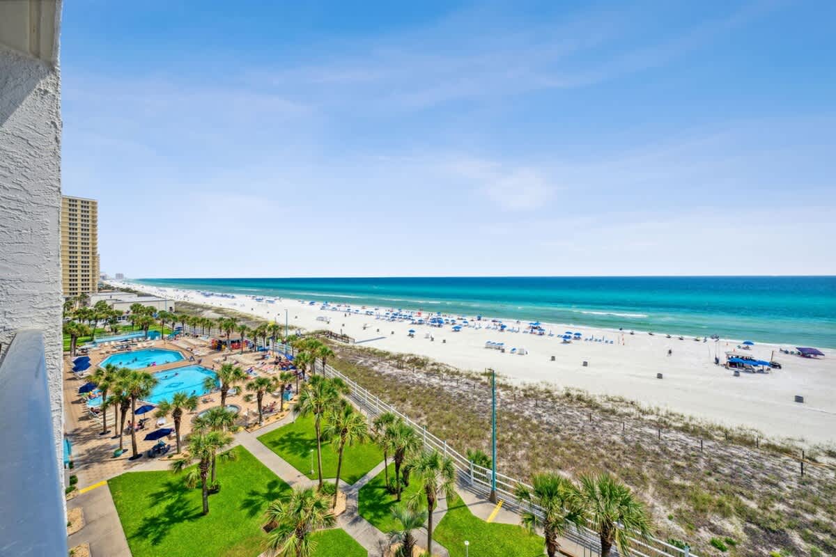 The Summit 702 – View of the beach and pool area from a balcony, featuring palm trees and blue umbrellas.