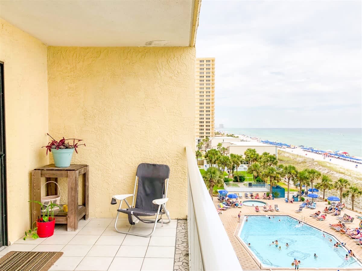 The Summit 611 – Balcony view overlooking beach, pool, and palm trees with a chair and wooden table.