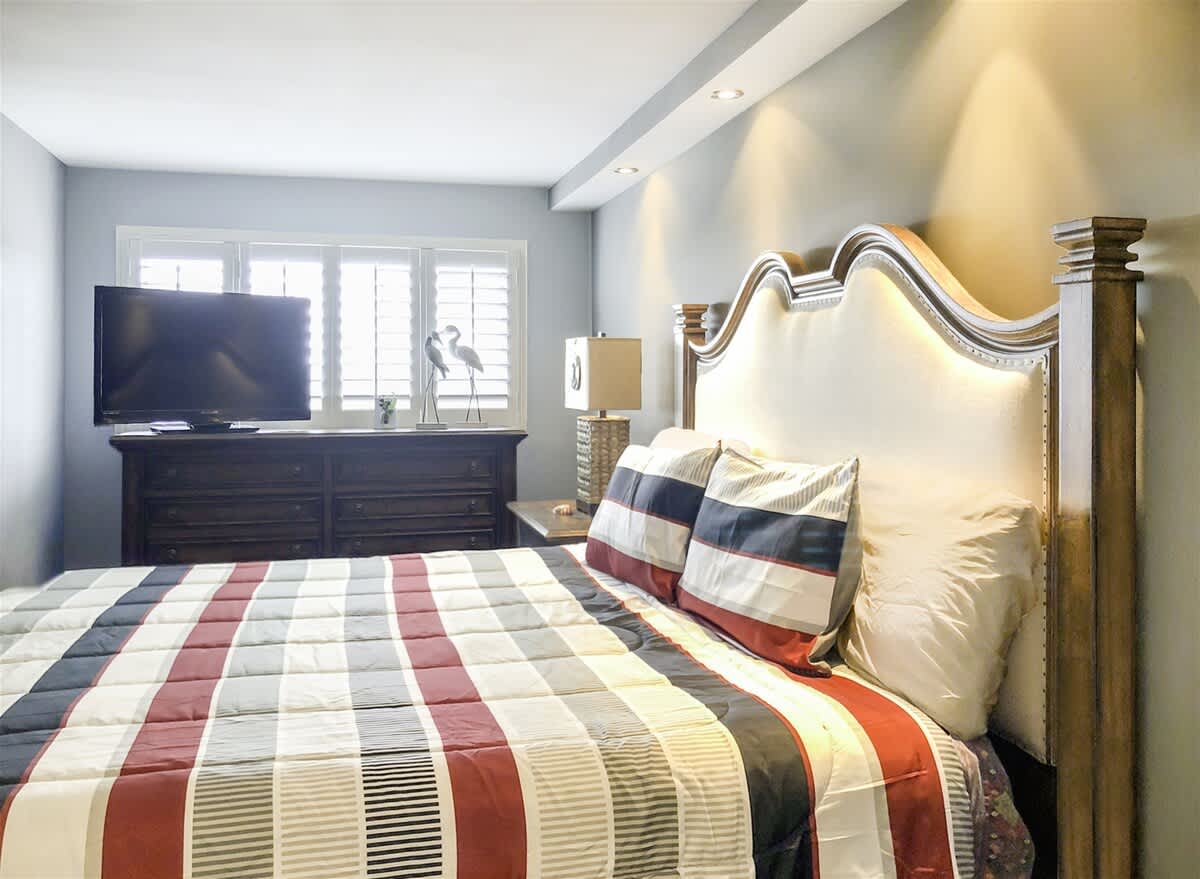 The Summit 611 – Bedroom with a striped comforter, a TV, dresser, and natural light from window shutters.