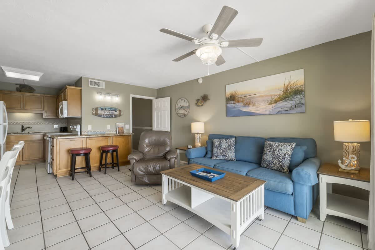 The Summit 1128 – Living room with blue sofa, brown armchair, coffee table, kitchen area, and beach-themed decor.