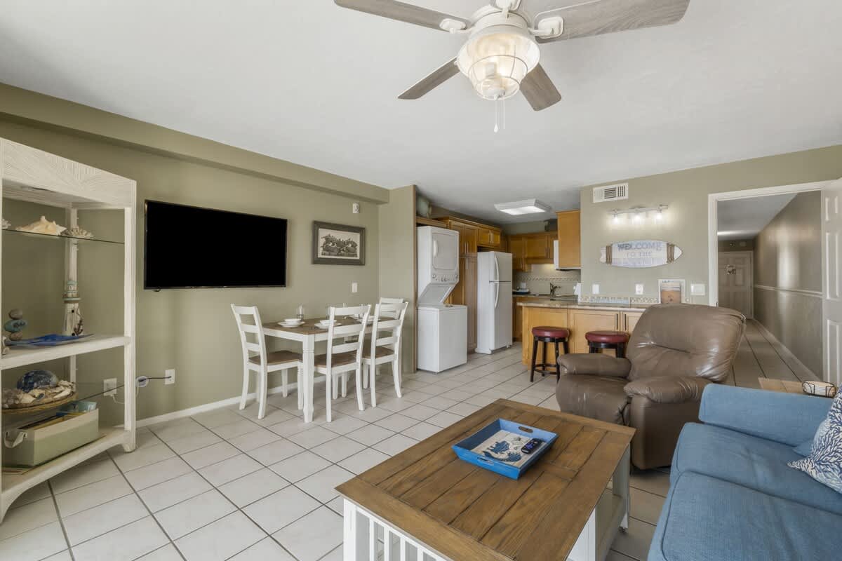 The Summit 1128 – Living room with a sofa, armchair, dining table, kitchen area, and wall-mounted TV.