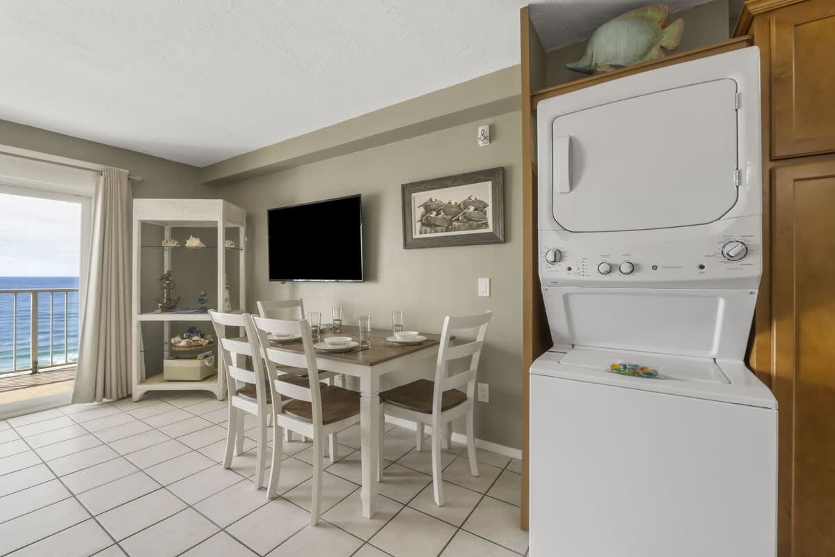 The Summit 1128 – Dining area with a table, chairs, wall-mounted TV, and laundry unit; ocean view through sliding glass doors.