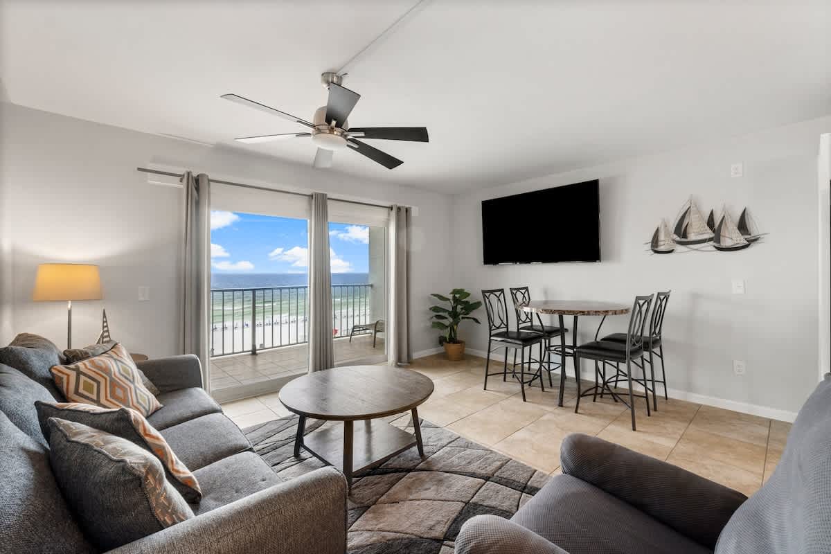 The Summit 620 – Living room with sofa, dining table, large window view of the beach, and a wall-mounted TV.