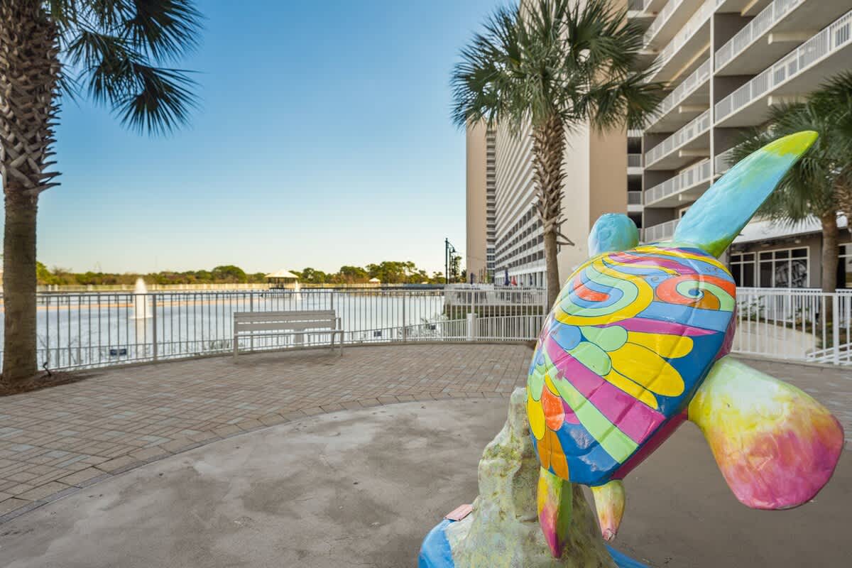 Laketown Wharf 1331 – Colorful turtle sculpture near water with palm trees and a building in the background.