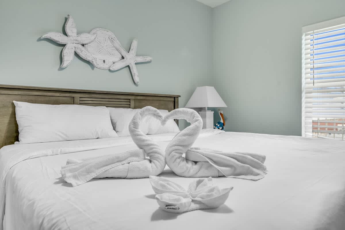 Laketown Wharf 1331 – Bedroom with a bed, towel swans, and blue decor near a window with blinds.