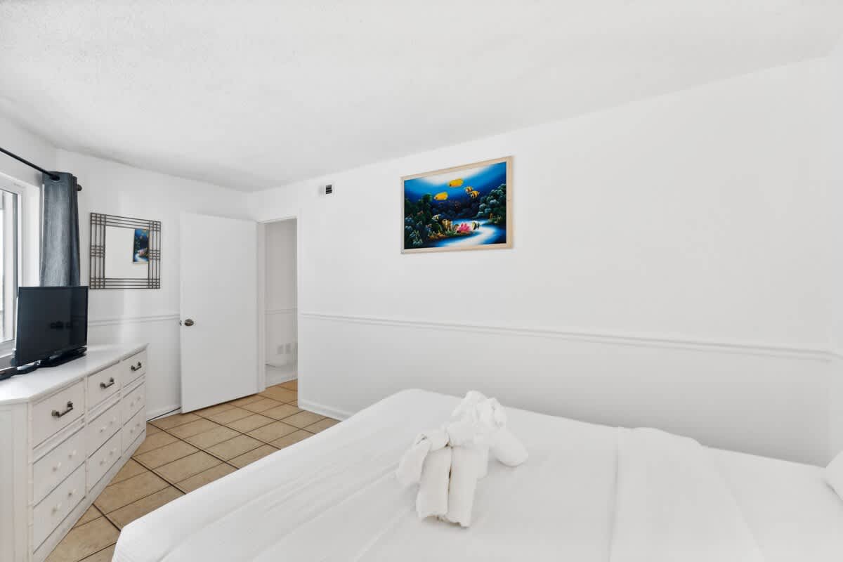 Moondrifter 608 – Bright bedroom with a bed, dresser, TV, artwork, and a door leading to another room.