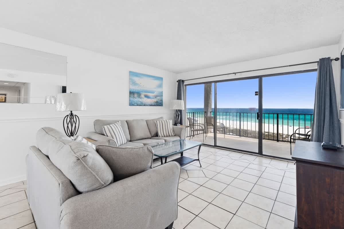 Moondrifter 608 – Living room with grey sofas, glass table, ocean view through sliding doors, and tile flooring.