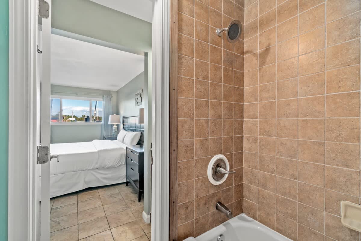 The Summit 105 – Bathroom view with tiled shower and adjoining bedroom featuring a bed, lamp, and window with a view.