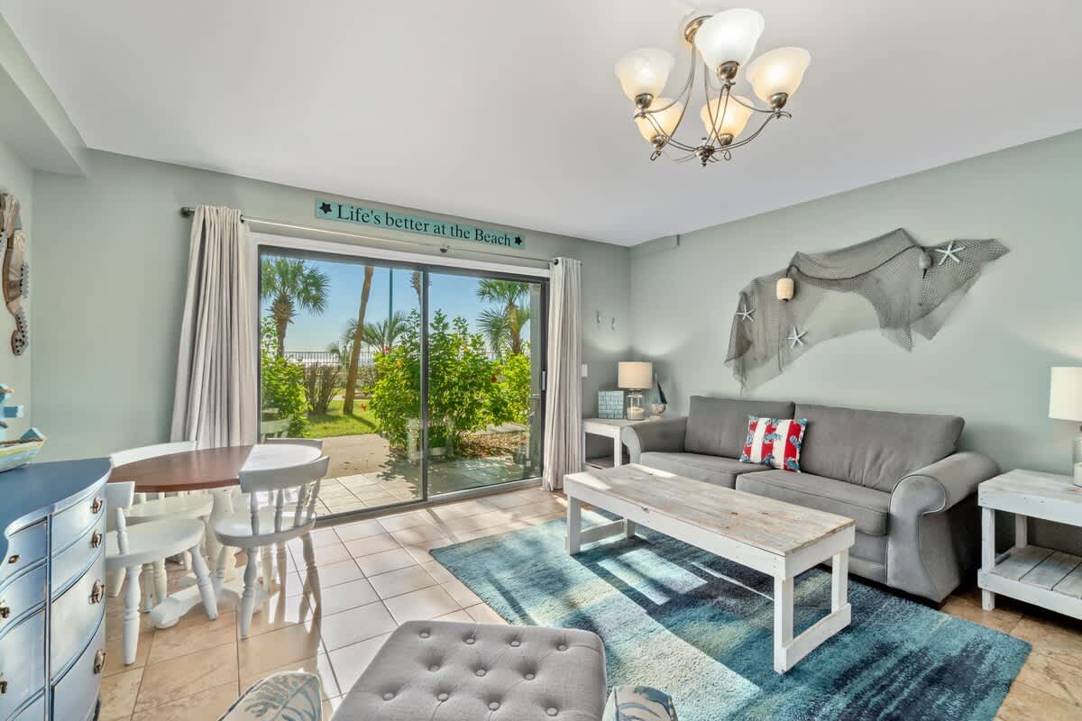 The Summit 105 – Living room with gray sofa, dining table, and view of palm trees through sliding glass doors.