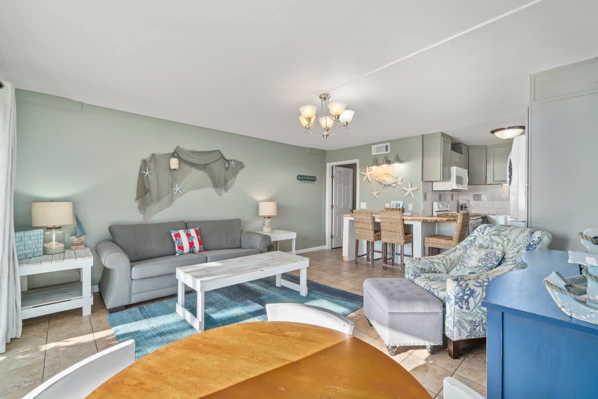 The Summit 105 – Living room with gray couch, dining table, and kitchen bar with stools, decorated in beach theme.