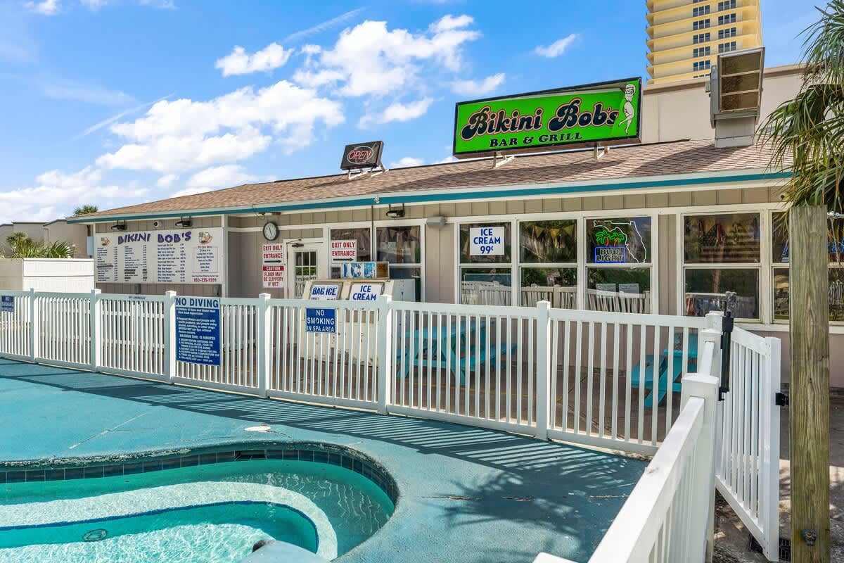 The Summit 228 – Outdoor bar and grill with pool area, white fencing, and blue seating under a clear sky.