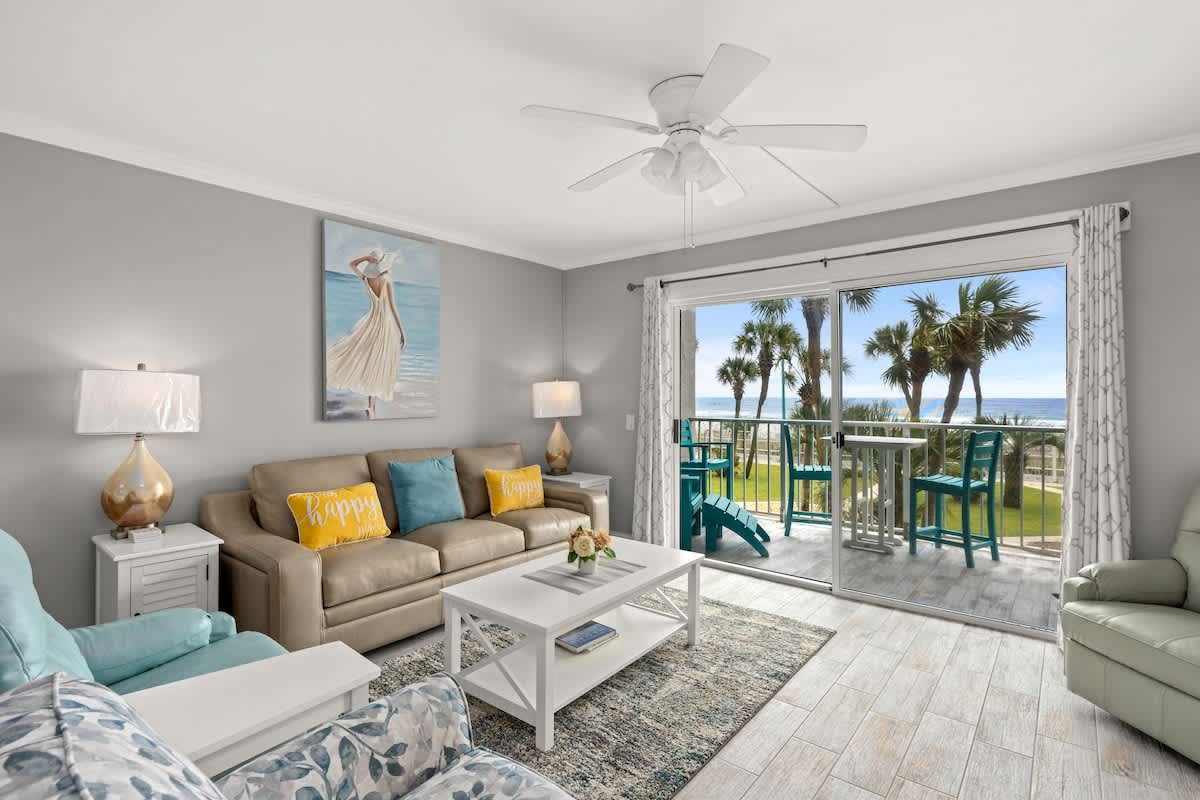 The Summit 228 – Living room with beige sofa, blue chairs, sliding glass doors, and ocean view with palm trees.