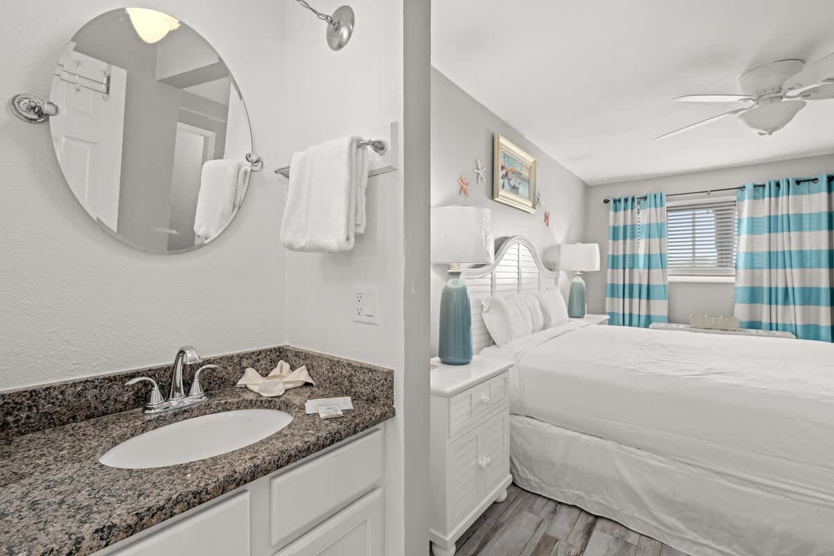 The Summit 228 – Bedroom with white bedding, striped curtains, large mirror, and a granite countertop sink.
