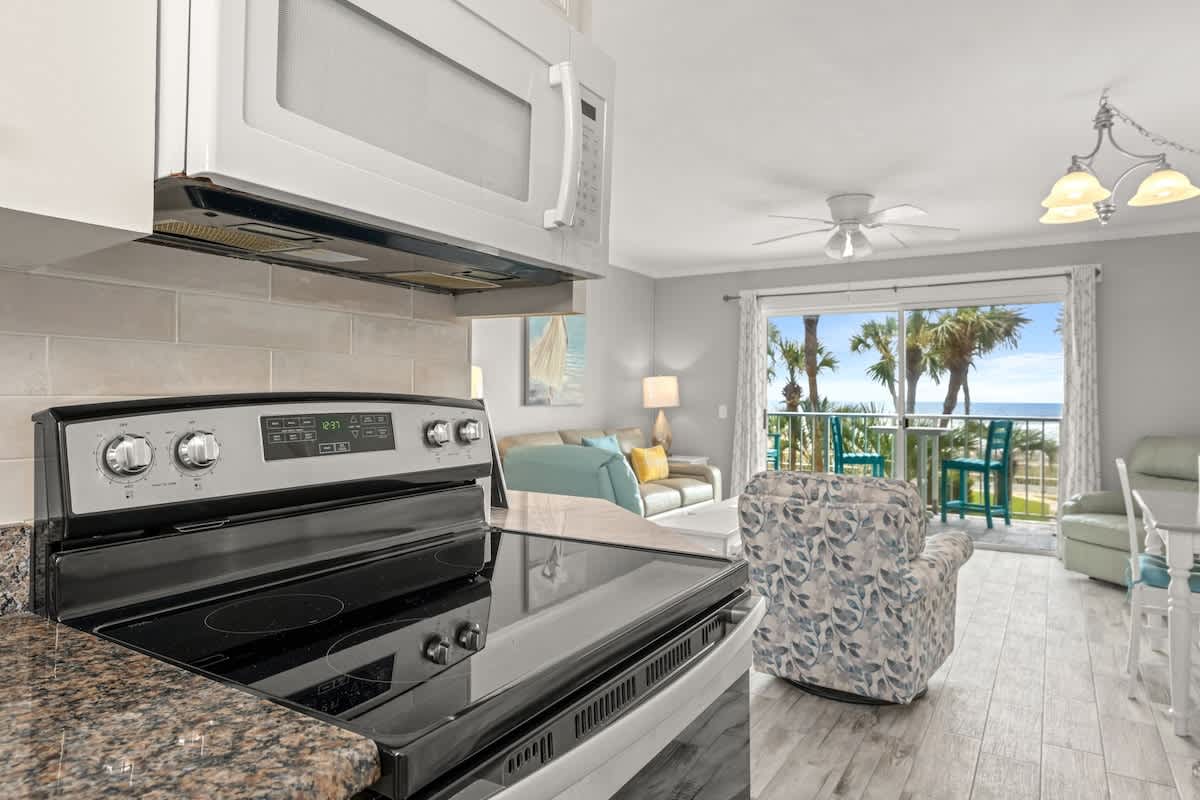 The Summit 228 – Kitchen with stove and microwave, living area with seating, balcony view of palm trees and ocean.