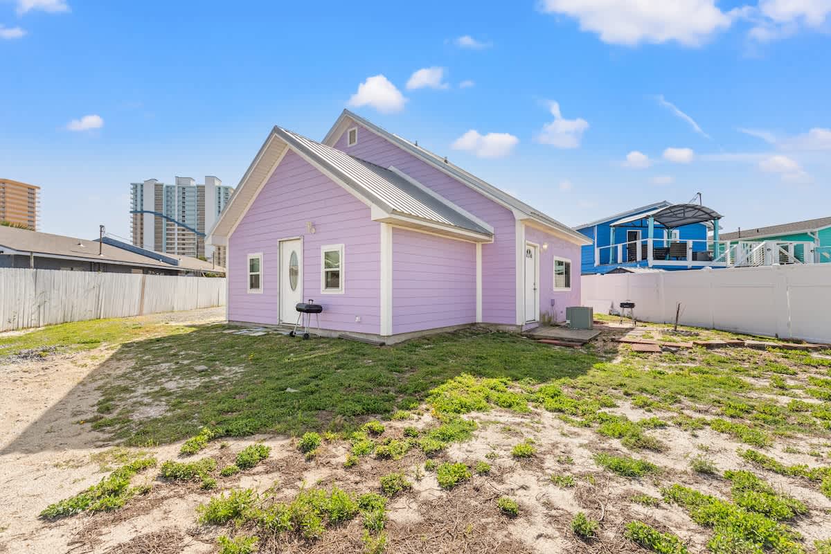 5218 Beach Dr C – Side view of a pink vacation rental cottage with grass and nearby buildings in the background.