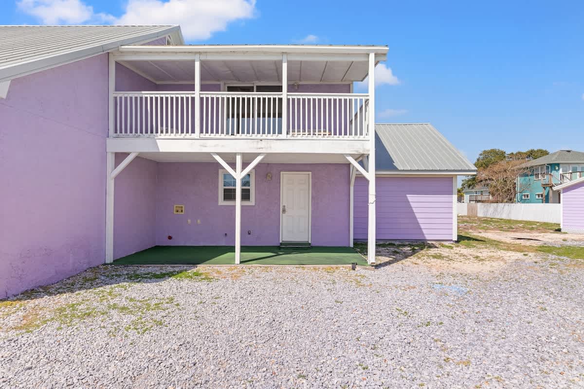 5218 Beach Dr C – Two-story, pastel purple vacation rental with a balcony, gravel yard, and green entryway.
