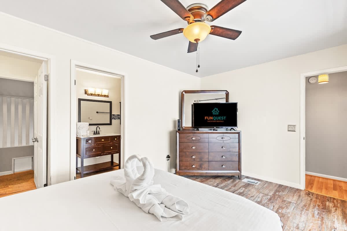 5218 Beach Dr C – Bright bedroom with ceiling fan, dresser, TV, and bathroom entrance visible. White bedding and towel swans on bed.