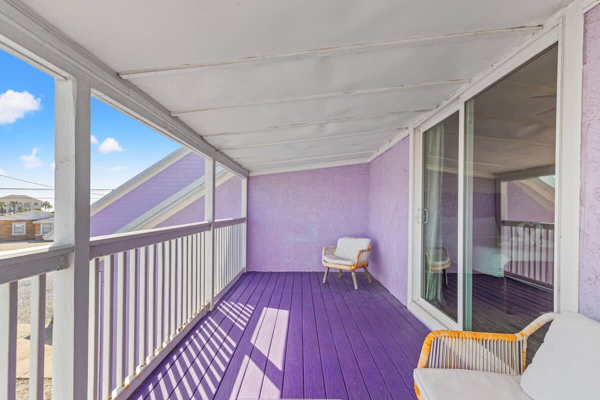 5218 Beach Dr C – Covered balcony with purple flooring, railing, and two seating areas, overlooking a distant house and blue sky.