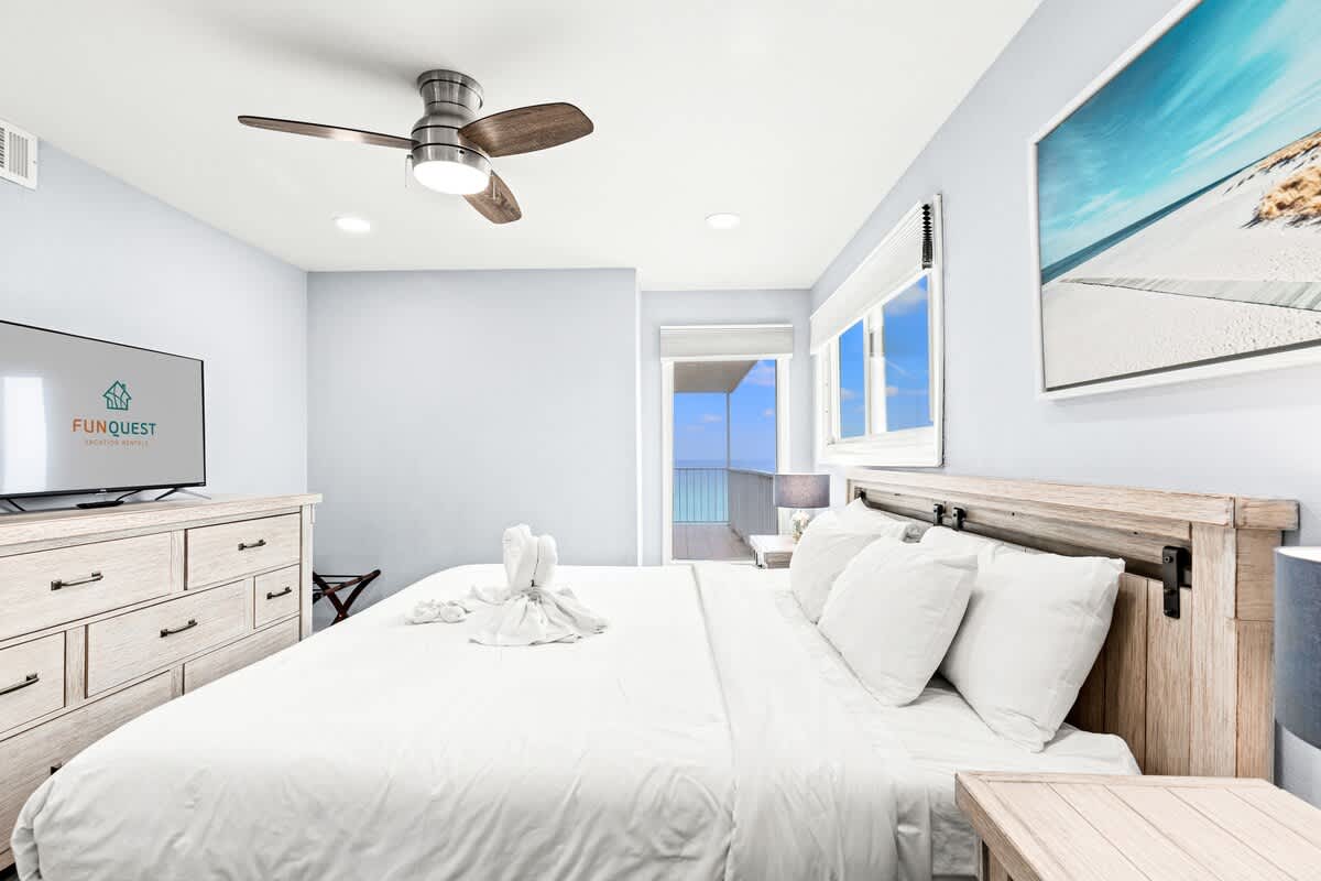 The Summit 701 – Cozy bedroom with white bedding, ceiling fan, TV, and sliding doors leading to a balcony view.