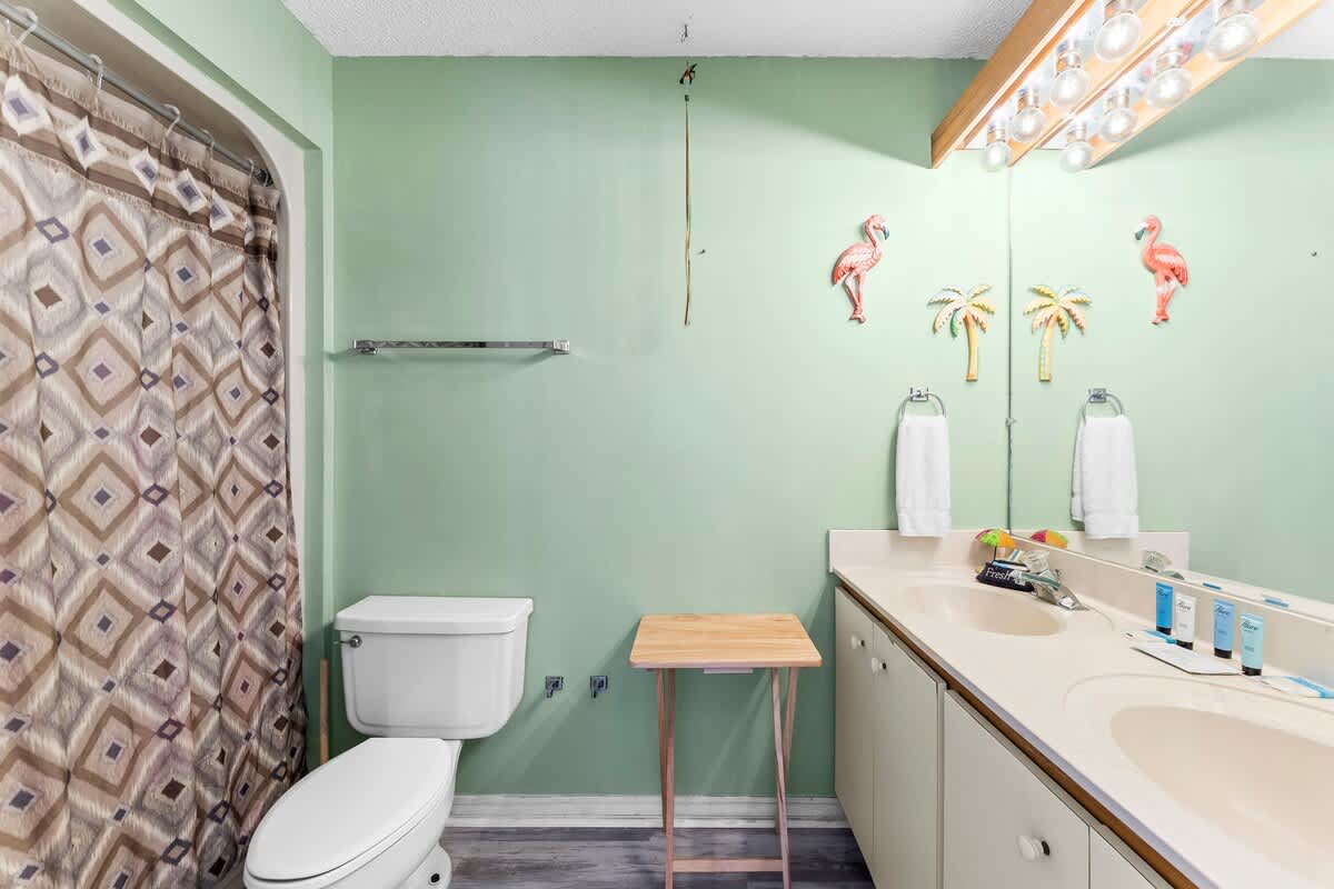 St Thomas SQ 1302B – Bathroom with green walls, double sink, toilet, shower curtain, and decorative flamingo and palm tree accents.