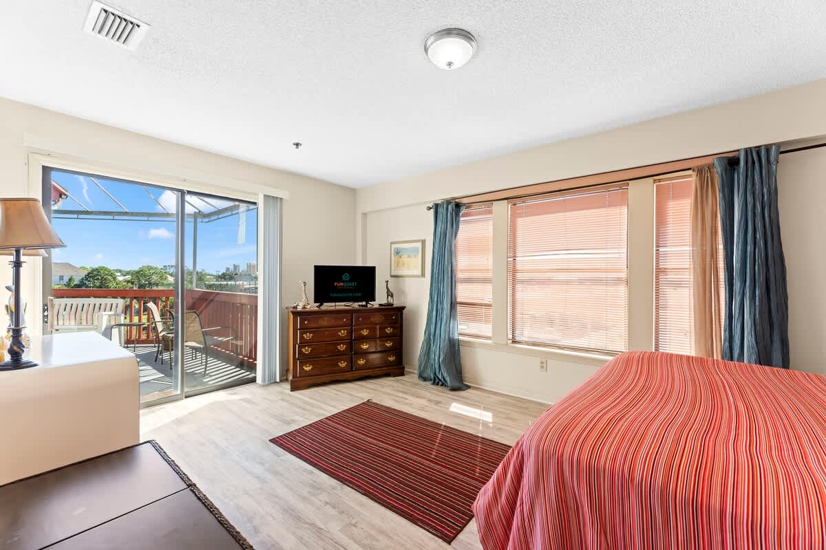 St Thomas SQ 1302B – Bright bedroom with a red-striped bedspread, dresser, TV, and sliding glass door to a balcony.