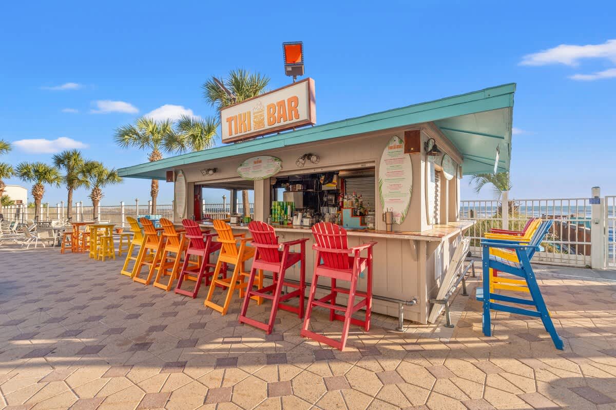 The Summit 1011 – Tiki bar with colorful chairs and palm trees, set against a clear blue sky.
