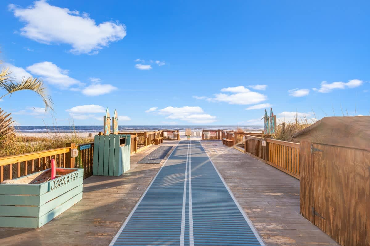 The Summit 1011 – Boardwalk leading to the beach with blue sky, palm trees, and toy-sharing boxes.