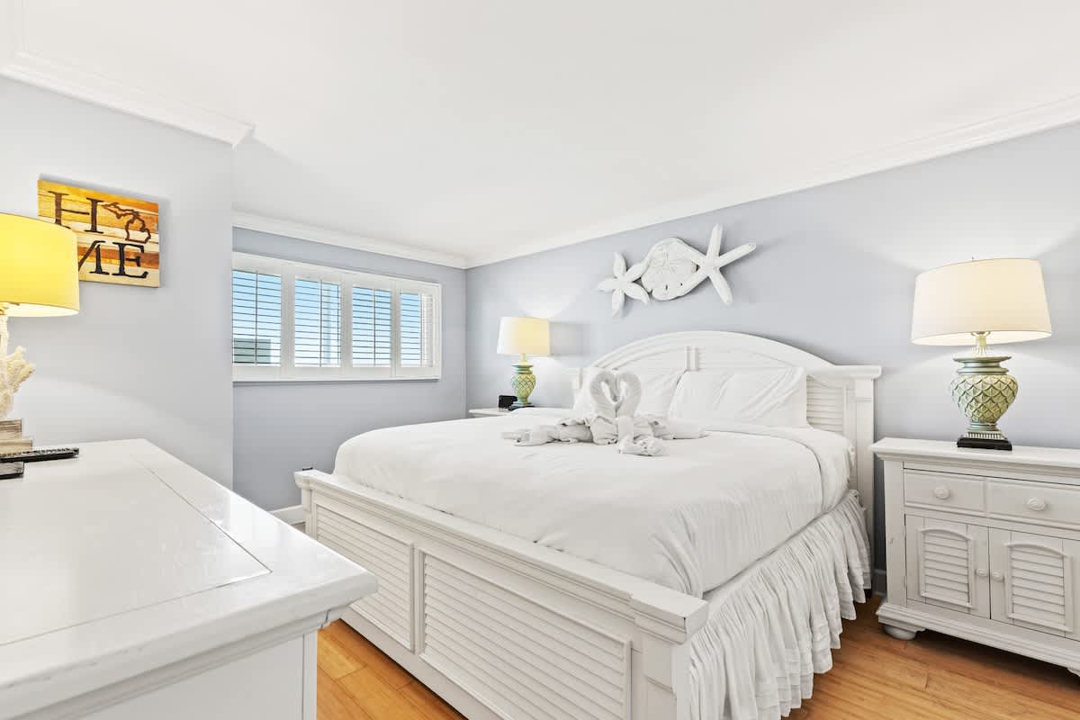 The Summit 1121 – Bright bedroom with a white bed, decorative lamps, and a window with shutters.